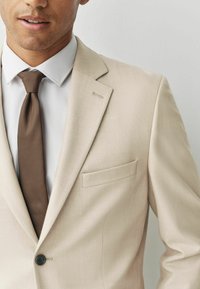 Beige blazer met een gestructureerd ontwerp, enkele knoop en subtiele textuur. Gepaard met een wit shirt en een bruine gestreepte stropdas.