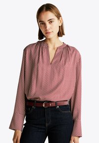 Rød mønstret bluse med lange ærmer, V-hals, subtile plisseringer, parret med et mørkt denimbælte med en metallisk spænde.