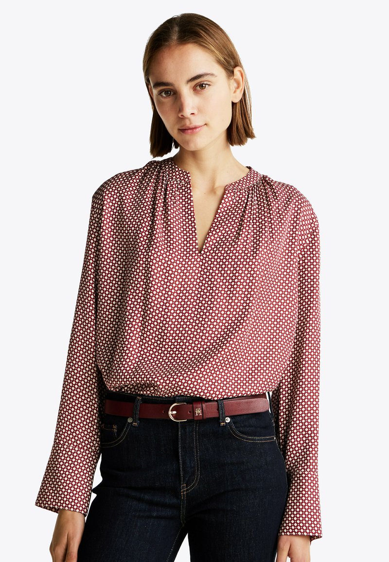 Rød mønstret bluse med lange ærmer, V-hals, subtile plisseringer, parret med et mørkt denimbælte med en metallisk spænde.