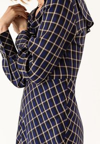 Kobieta ubrana w granatową sukienkę w beżową kraciastą wzór, z długimi rękawami i wcięciem w talii, delikatnie dotykająca twarzy.