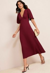 Abito bordeaux a lunghezza media con maniche corte, tessuto texturizzato e scollo a V. Abbinato a una pochette dorata e sandali neri con tacco.