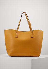 Grande borsa a tracolla in pelle giallo senape con superficie testurizzata e doppie spalline, esposta su un piedistallo bianco su uno sfondo chiaro.