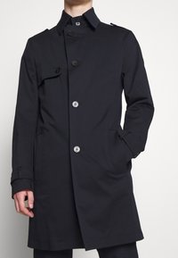 Mörk marinblå trenchcoat med dubbelknäppt design, stora knappar, klassisk krage och sidofickor. Slät, lättviktsmaterial.