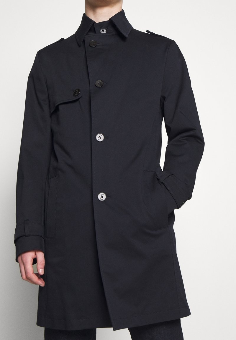Mörk marinblå trenchcoat med dubbelknäppt design, stora knappar, klassisk krage och sidofickor. Slät, lättviktsmaterial.