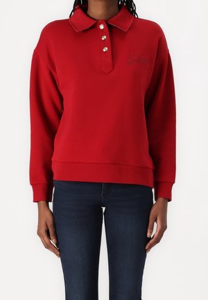 Personne portant un polo rouge à manches longues avec des boutons dorés et un jean bleu foncé sur un fond blanc uni.