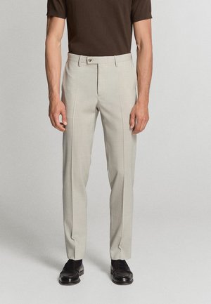Hombre con pantalones ajustados beige claro, una camisa marrón de manga corta y zapatos de vestir negros, de pie frente a un fondo liso.