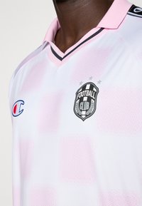 Zbliżenie osoby noszącej biało-różową koszulkę piłkarską w kratę z czarno-białym herbem klubu piłkarskiego i logo Champion.