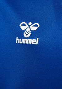 Blauer Stoff mit glatter Textur und einem weißen Hummel-Logo, das einer stilisierten Biene ähnelt, auf der linken Seite platziert.