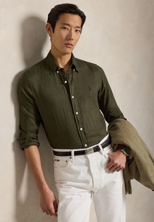 Muškarac u maslinasto zelenoj košulji s gumbima i bijelim hlačama drži smeđu jaknu, stoji uz bež zid.