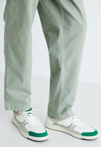 Sportowe sneakersy z białą skórzaną bazą, zielonymi akcentami i szarymi zamszowymi panelami. W połączeniu z luźnymi, jasnoszarymi spodniami.