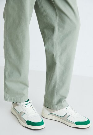 Sportliche Sneakers mit einem weißen Lederoberteil, grünen Akzenten und grauen Wildlederpaneelen. Kombiniert mit locker sitzenden hellgrünen Hosen.