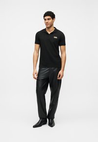 Άνδρας στέκεται φορώντας μαύρο πόλο με μικρό λογότυπο, μαύρο δερμάτινο παντελόνι και μαύρα μυτερά παπούτσια μπροστά σε λευκό φόντο.