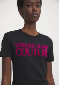 Svart kortermet t-skjorte med fet, rosa fløyelstekst som leser "VERSACE JEANS COUTURE." Enkelt design med klassisk rund hals.