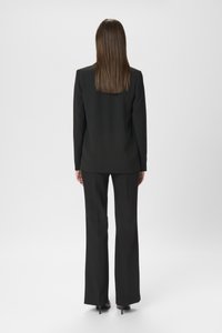 Giacca nera con vestibilità su misura, abbinata a pantaloni coordinati sempre con vestibilità su misura. Tessuto liscio e dettagli minimi. Tacchi visibili.