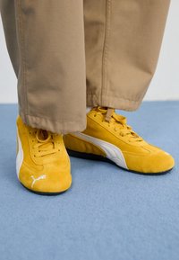 Gula mockasiner med vita detaljer, med en låg profil och texturerad yta, kombinerade med beige, lätt utsvängda byxor.