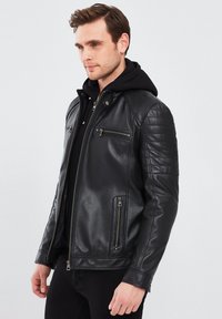 Chaqueta de cuero negra con un diseño acolchado en los hombros, bolsillos con cremallera y una sudadera con capucha negra debajo. Textura suave.