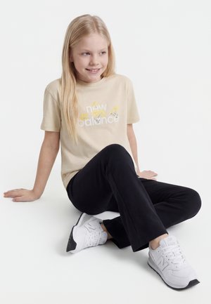 Ragazza bionda seduta a gambe incrociate, indossa una t-shirt beige New Balance, pantaloni neri e sneakers bianche New Balance su sfondo bianco.