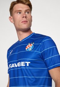 Maglia sportiva blu con strisce bianche orizzontali, caratterizzata da un logo sul petto e un design a scollo a V. Realizzata in un materiale traspirante e testurizzato.