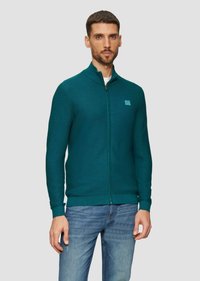Turquoise ritsvest gemaakt van gestructureerde stof, met een hoge kraag en een klein logo-opdruk op de borst. Gepaard met blauwe spijkerbroeken.