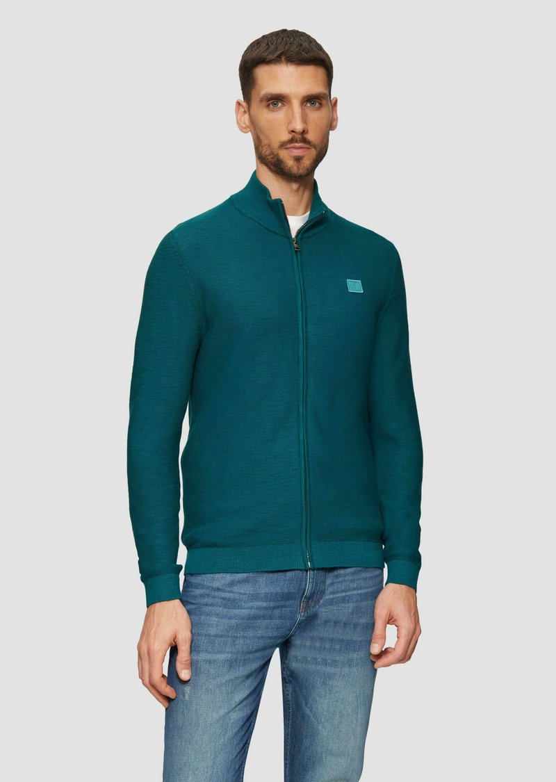 Turquoise ritsvest gemaakt van gestructureerde stof, met een hoge kraag en een klein logo-opdruk op de borst. Gepaard met blauwe spijkerbroeken.