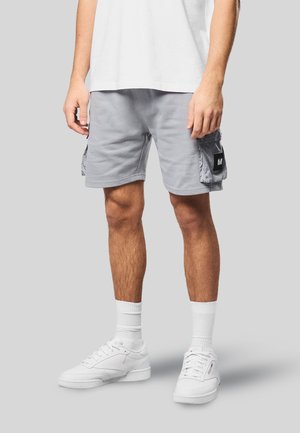 Mężczyzna ubrany w jasnoszare szorty typu cargo, białe skarpetki do kostek, białe trampki oraz białą koszulkę, stojący na szarym tle.