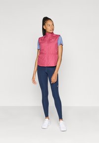 Könnyű, piros szigetelt mellény magas gallérral, cipzáros elülső résszel és rugalmas derékkal, kék rövid ujjú felsővel és sötétkék leggings-szel párosítva.