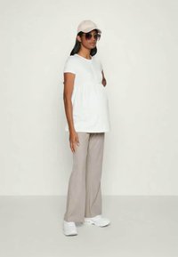 T-shirt de maternité blanc à manches courtes, pantalon large beige, baskets blanches, casquette rose et lunettes de soleil noires. Design simple avec une coupe décontractée.