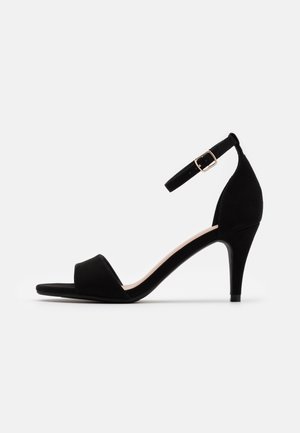 Sandale à talon haut en daim noir avec une bride avant unique, une bride de cheville avec une boucle dorée, et un design à bout pointu.