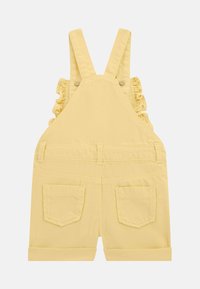 Pantalones de mezclilla amarillos con tirantes con volantes, bolsillo frontal y dos bolsillos traseros. Dobladillos enrollados en las aperturas de las piernas para un ajuste informal.