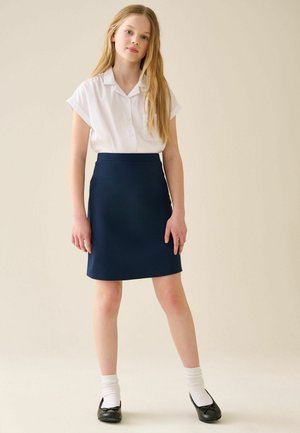Młoda dziewczyna z długimi blond włosami, ubrana w białą bluzkę z krótkim rękawem, granatową spódnicę, białe skarpetki i czarne baleriny, stojąca na jednolitym tle.