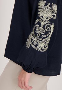 Marineblauwe blouse met lange, opgeblazen mouwen, voorzien van ingewikkeld lichtgroen borduurwerk in bloem- en spiraalpatronen op de linkermouw.