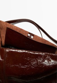 Brązowa torba typu tote ze skórzaną, błyszczącą powierzchnią zewnętrzną i miękkim, zamszowym wnętrzem. Posiada ciemnobrązowy skórzany akcent oraz regulowane paski.