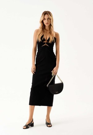 Black knit dress with a fitted silhouette and a twist front detail. Paired with a black textured handbag and strappy block-heeled shoes.