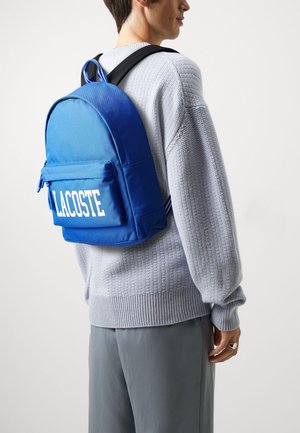 Blauwe Lacoste rugzak met voorzak met rits en wit logo, gemaakt van duurzaam materiaal, voorzien van zwarte verstelbare banden en een handvat aan de bovenkant.