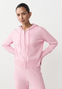 Różowa bluza z kapturem zapinana na zamek, z prążkowanym obszyciem na dole i rękawach, dwoma przednimi kieszeniami oraz kapturem ze ściągaczem. Wykonana z miękkiego materiału o gładkiej fakturze.