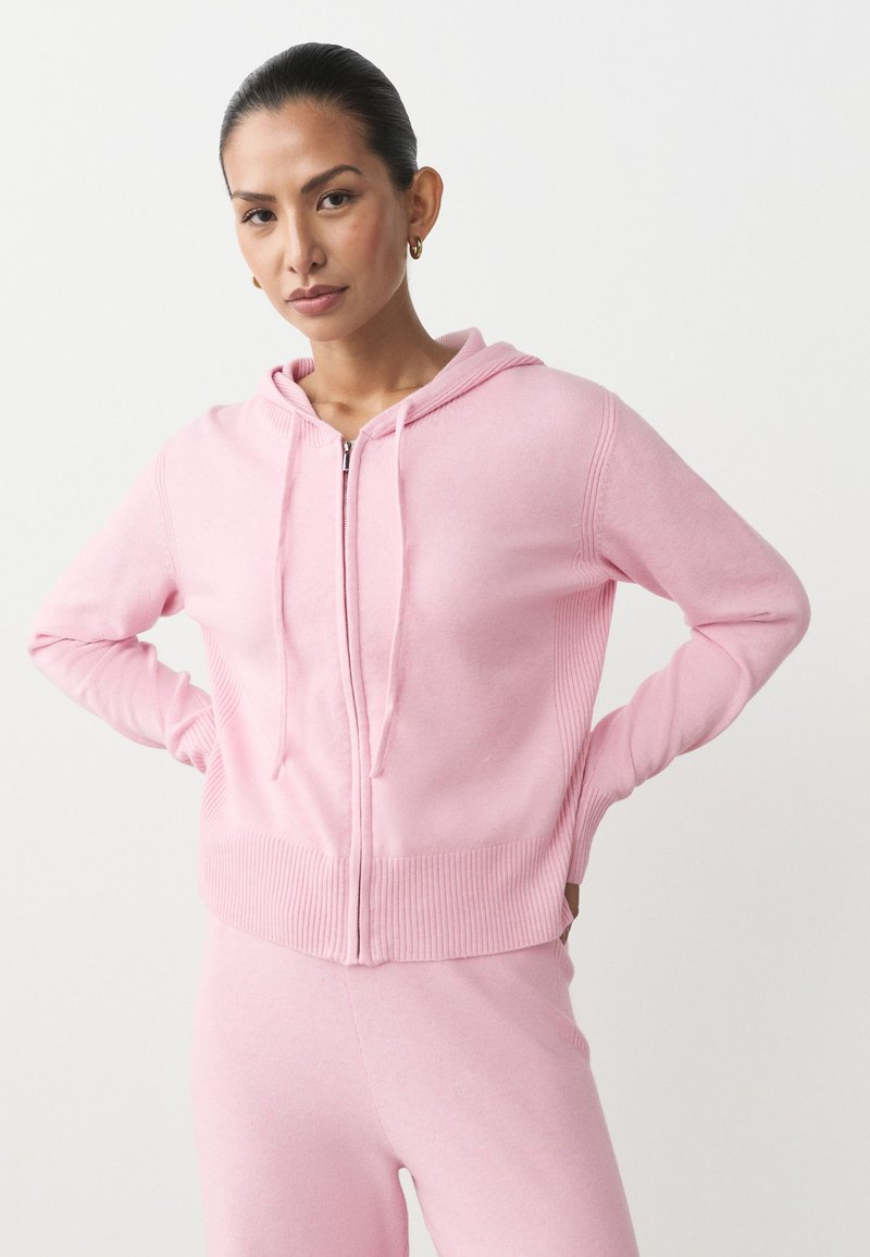 Różowa bluza z kapturem zapinana na zamek, z prążkowanym obszyciem na dole i rękawach, dwoma przednimi kieszeniami oraz kapturem ze ściągaczem. Wykonana z miękkiego materiału o gładkiej fakturze.