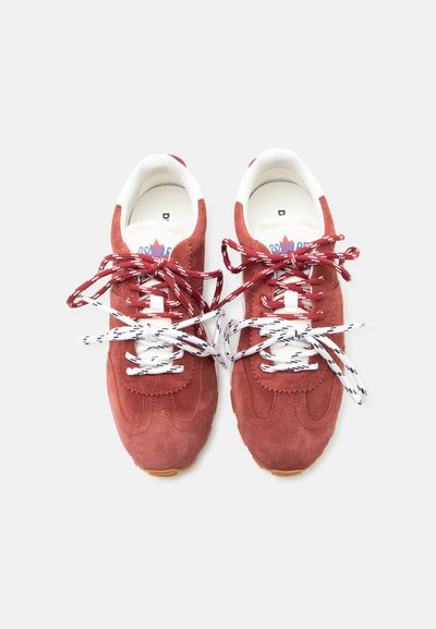 Paire de baskets en daim rouge avec collerettes blanches rembourrées, lacets blancs et rouges à motif noués en nœuds sur des lacets blancs et noirs, sur fond blanc.