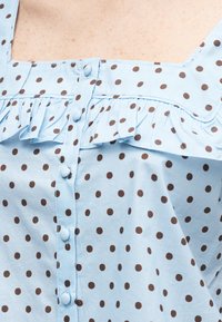 Ανοιχτό μπλε μπλούζα με καφέ πουά, κουμπιά καλυμμένα με ύφασμα, λεπτομέρεια με βολάν και φαρδιές τιράντες.