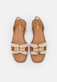 Sandales plates marron avec deux brides tissées beige comportant une boucle décorative dorée. Semelle intérieure lisse avec un design minimaliste.