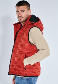 Rote gesteppte Steppweste mit Kapuze und strukturierter Oberfläche sowie Seitentaschen, getragen über einem beige gerippten Langarmshirt. Sichtbarer Reißverschluss.