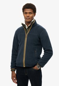 Chaqueta de forro polar azul marino con un cierre dorado en la parte delantera, bolsillos laterales y cuello elevado, superpuesta sobre un suéter verde texturizado y combinada con jeans oscuros.