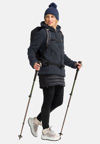 Fleece jack in donkerblauw, geïsoleerde rok, zwarte leggings en lichte schoenen met multikleurige accenten. Rugzak en wandelstokken inbegrepen.