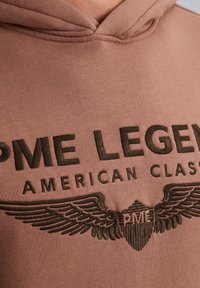 Hoodie marron avec un logo sur le devant contenant le texte brodé "PME LEGEND AMERICAN CLASSIC" et un design de bouclier ailé en marron foncé.