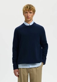 Pull en laine marine foncé avec un col rond et des poignets côtelés, superposé à une chemise rayée bleu clair, accompagné de pantalons beiges.