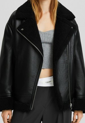 Person wearing a black shearling-lined biker jacket over a gray ribbed crop top and black pants with a visible white waistband label.