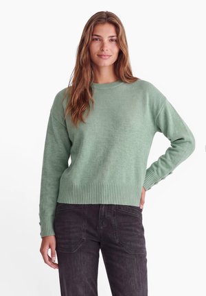 Donna con capelli castani lunghi che indossa un maglione a coste verde chiaro e jeans neri, in piedi con una mano sui fianchi davanti a uno sfondo bianco.