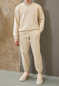 Sudadera beige de cuello redondo con puños acanalados, combinada con pantalones de chándal ajustados a juego. Llevada con zapatillas blancas con detalles en naranja.