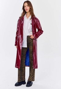 Burgundské lakované dlhé kabát overený na bielej košeli, kombinovaný s nohavicami s leopardím vzorom a čiernymi platformovými topánkami. Výrazný lesk a farebný kontrast.