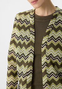 Sweter kardigan w oliwkowej zieleni, z wzorem w zygzaki w kolorach czarnym, białym i jasnoróżowym. Teksturowany materiał z otwartym przodem.