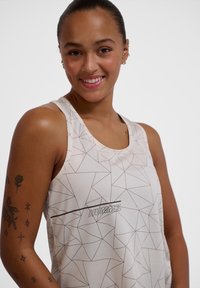 Svetlo béžový tank top vyrobený z textúrovanej látky s geometrickým vzorom v tmavšej hnedej farbe. Na spodnej strane sa nachádza logo "HUMMEL".